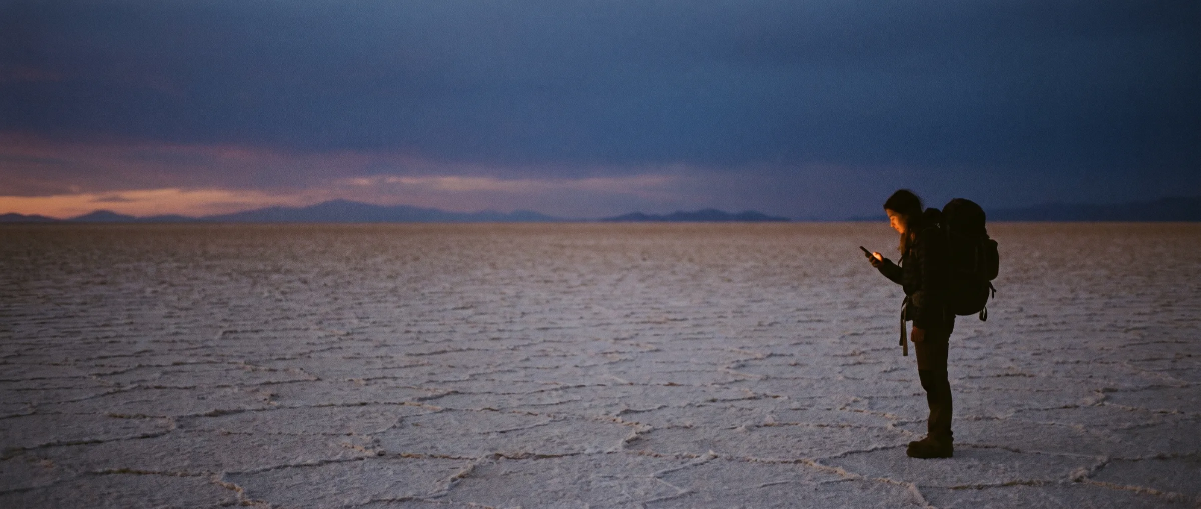 A lone traveller on a vast salt flat at dusk, lit by a phone screen